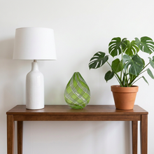 Load image into Gallery viewer, Computer-generated illustration: Jeffrey P'an green and clear teardrop art glass vase on a wooden console table beside a white lamp and a potted plant