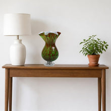 Load image into Gallery viewer, Computer-generated illustration: green and brown splatter glass vase with ruffled rim and clear foot on a wooden console table beside a white lamp and a small potted plant
