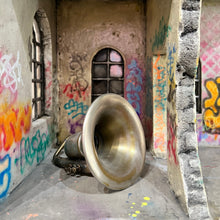 Load image into Gallery viewer, Close-up of interior room with brass horn lying on the floor, surrounded by graffiti-covered walls and arched window.
